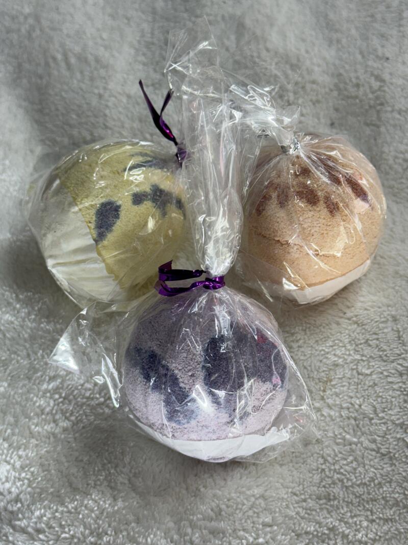 Three bath bombs individually wrapped in clear plastic are arranged on a white, textured surface. The bombs are light green with dark green spots, peach with orange spots, and purple with dark purple spots, tied with purple and silver ribbons.