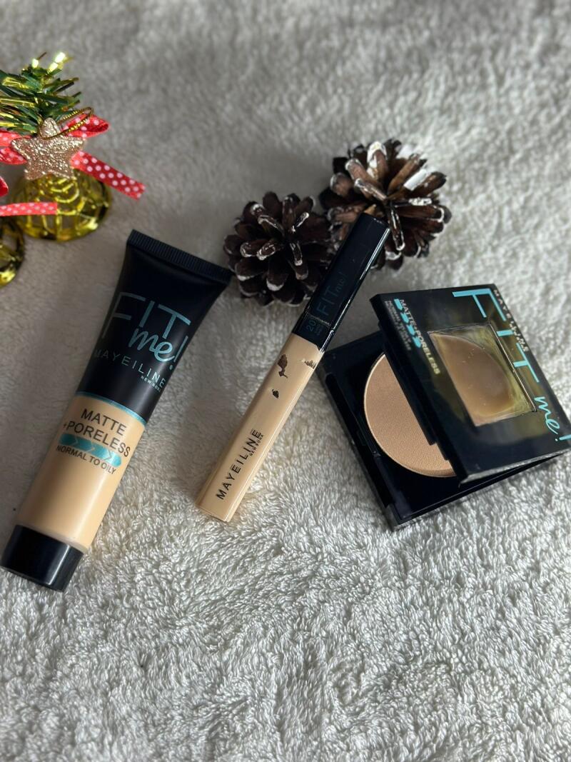 The image shows a collection of Maybelline Fit Me! makeup products including foundation, concealer, and powder, arranged on a textured white surface, alongside two pine cones and a small decorative Christmas tree.