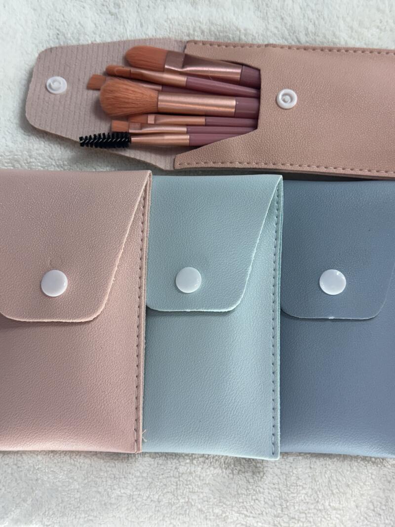 The image shows three closed pouches in pink, light blue, and gray, each with a white snap closure, along with an open pouch containing several makeup brushes with pink handles and copper ferrules.