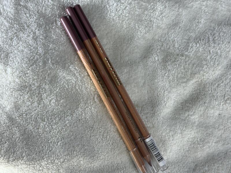 Three lip liners with brown wooden barrels and maroon ends are lying on a white towel.