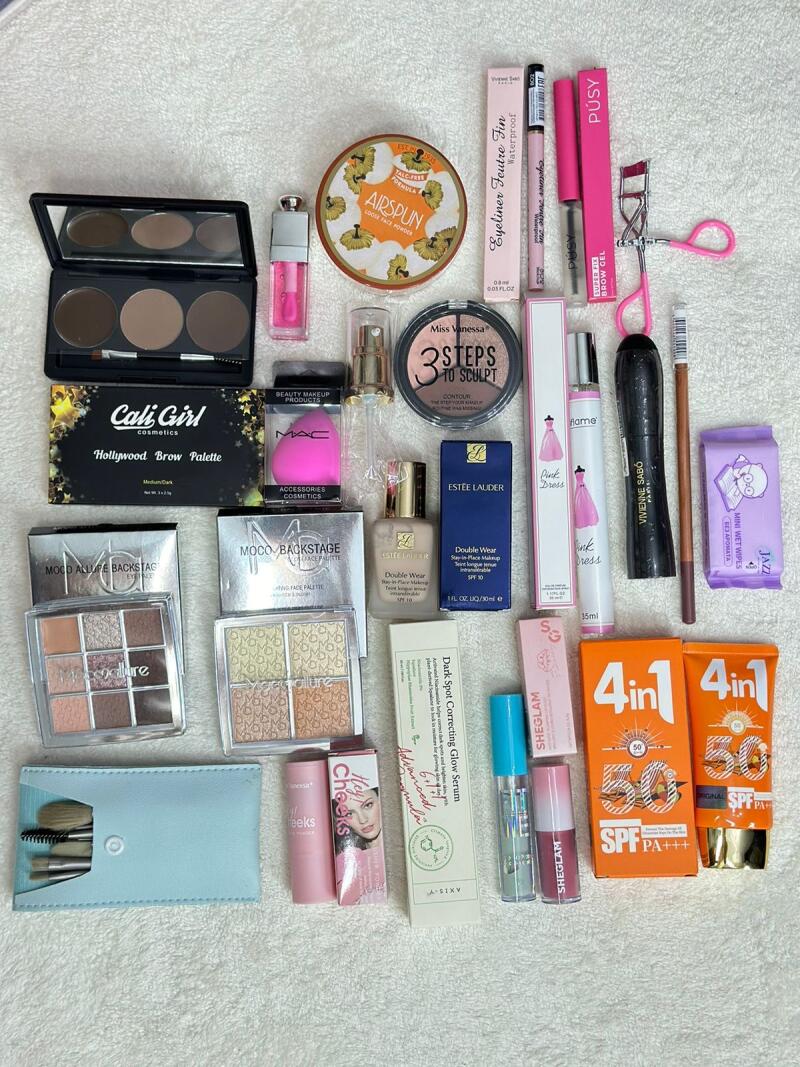 The image shows a collection of various makeup and cosmetic products arranged on a white surface, including eyeshadow palettes, brow kits, foundation, serum, sunscreen, mascara, eyeliner, lip gloss, and makeup brushes.