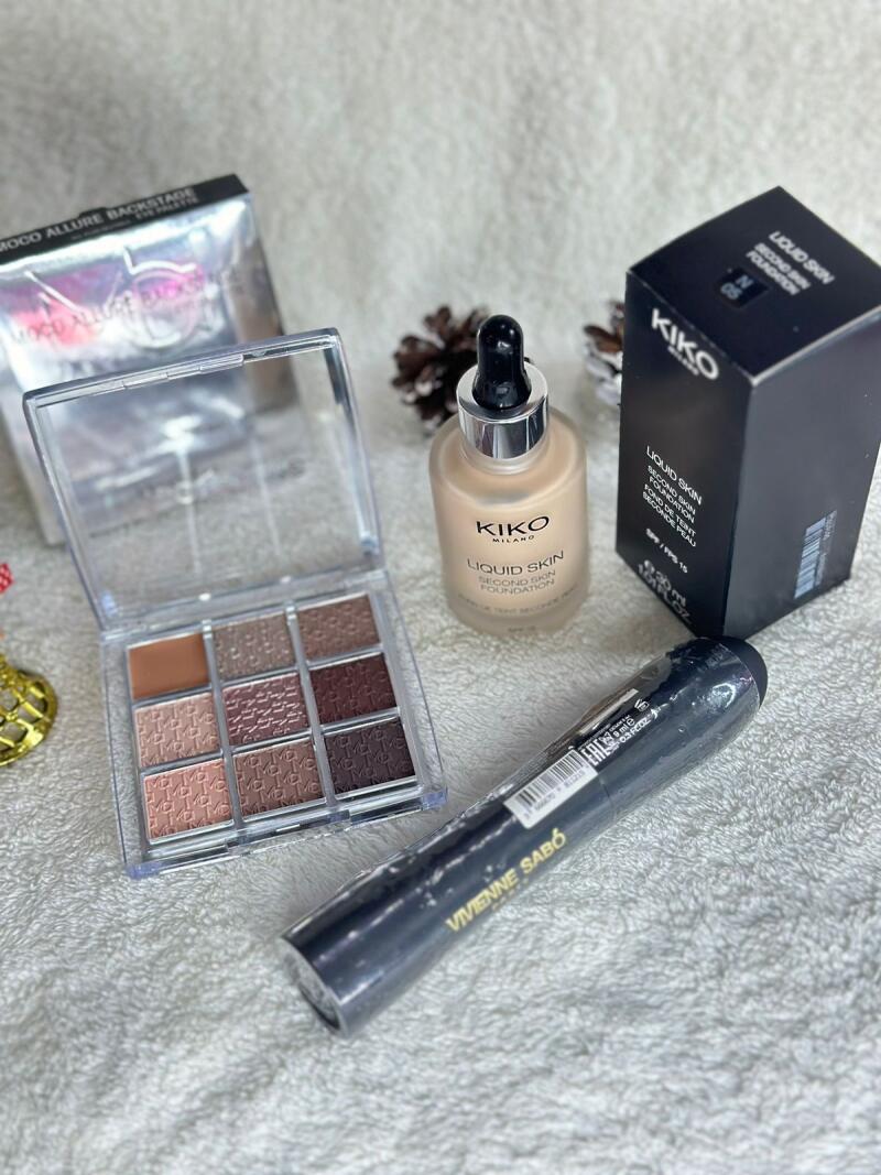 The image shows a collection of makeup products including a Kiko Milano eyeshadow palette, Kiko Milano liquid skin foundation with box, and Vivienne Sabó mascara, all arranged on a white textured surface.
