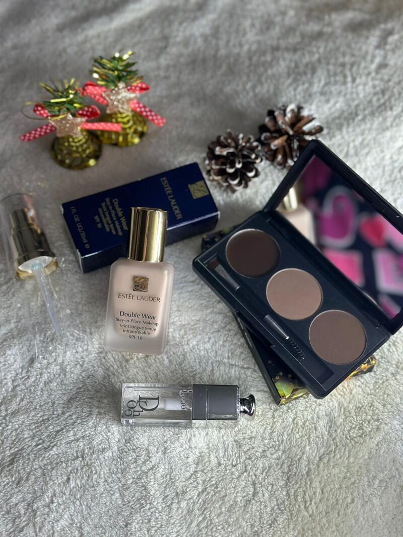 The image displays an assortment of beauty products including Estee Lauder Double Wear foundation, a Dior lip gloss, and a brow palette with a mirror, along with Christmas decorations such as pine cones and small golden bells.