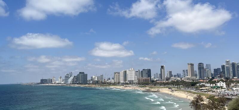 The image shows a coastal cityscape under a partly cloudy sky, with a variety of modern buildings and skyscrapers along the shoreline.