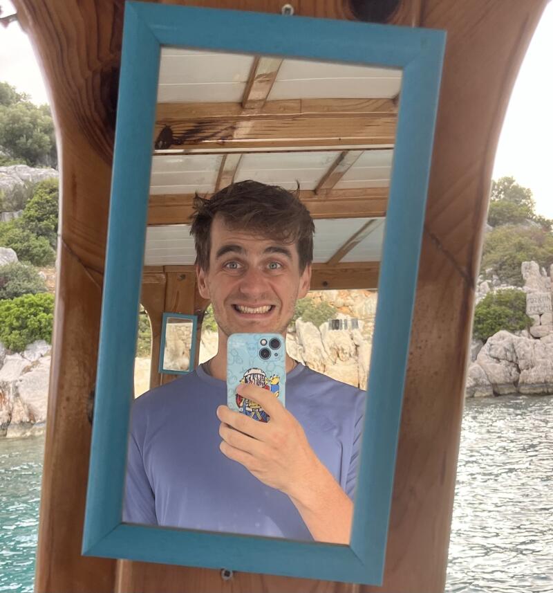A man with brown hair is taking a selfie in a blue-framed mirror, wearing a blue shirt and holding a phone with a cartoon case, with a coastal landscape visible in the background.