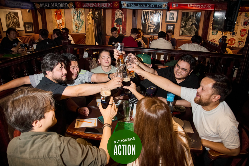 A group of people are toasting with glasses of beer at a table in a pub, under sports memorabilia, with the text 'КИНОКВИЗ ACTION'.