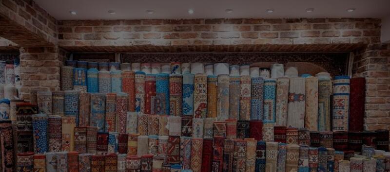 The image shows a large collection of rolled-up carpets and rugs with various colors and patterns, stacked densely inside a shop with brick walls.