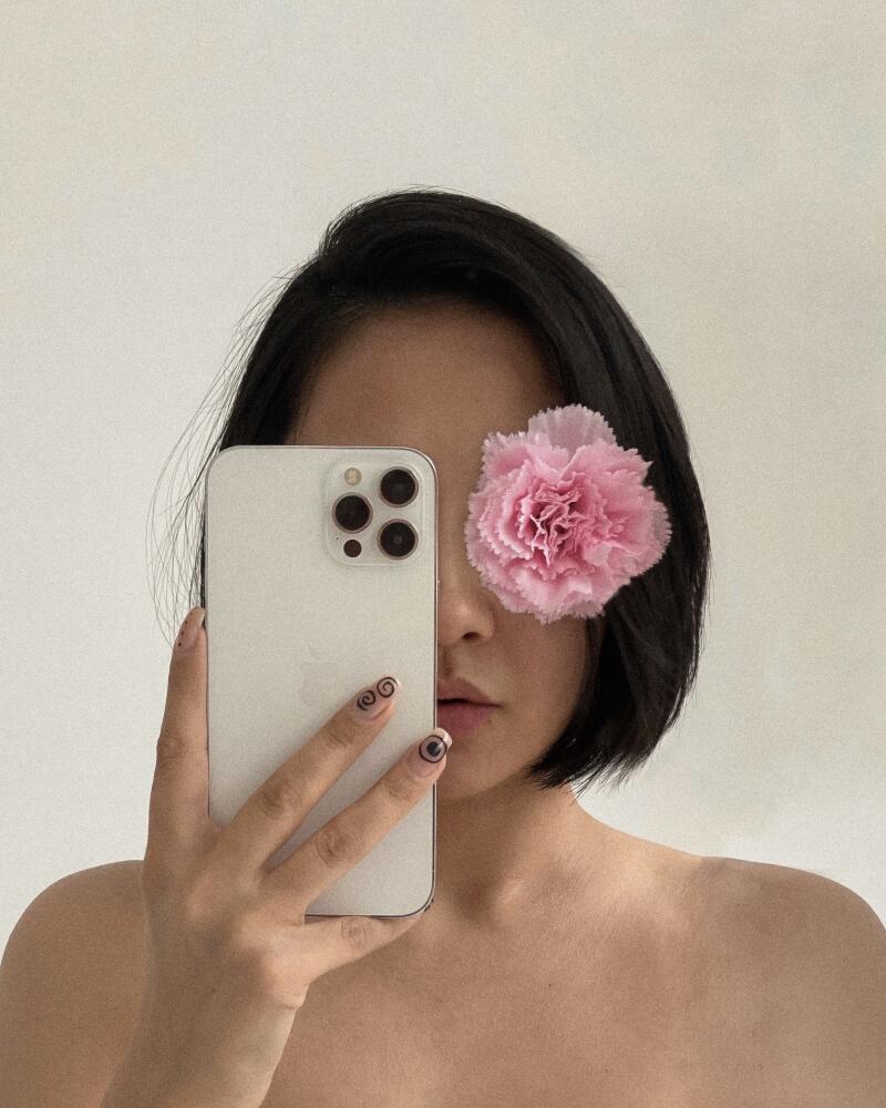A person with short black hair holds a white smartphone up to their face, partially obscuring it, with a pink flower covering one eye.