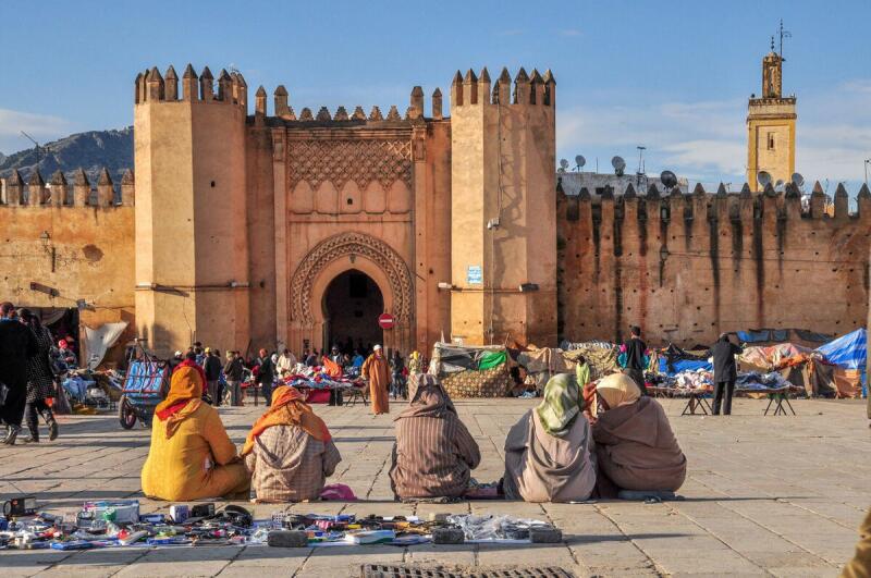 A bustling outdoor market scene features several people sitting on the ground in the foreground, displaying their wares for sale, with an ancient gate and city wall in the background under a clear sky.