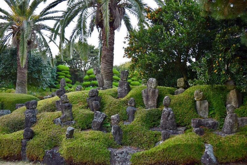 The image shows a garden with stone statues and palm trees, surrounded by green bushes and trees.