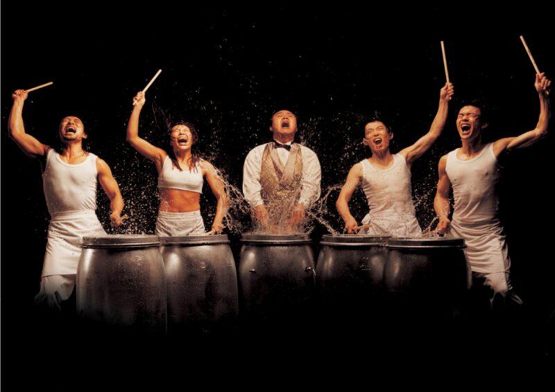 Five people are enthusiastically drumming on metal barrels, with water splashing around them, set against a dark background.