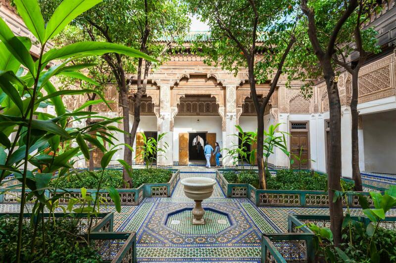 The photo shows a courtyard with green plants, trees, a fountain, and people walking through an open doorway in the background.