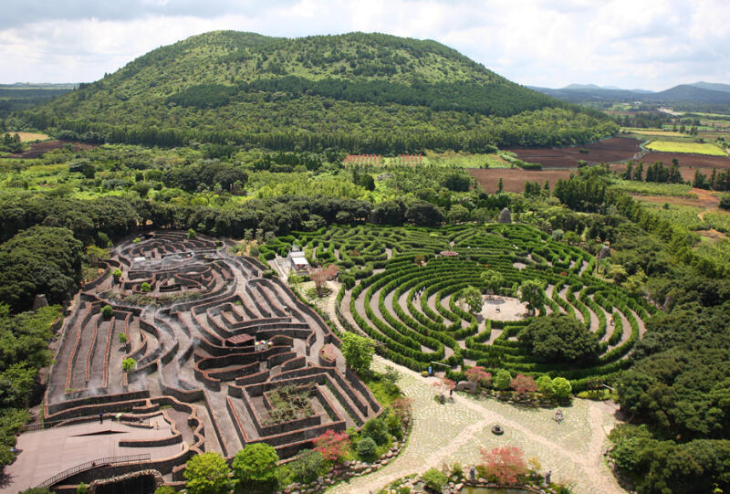 An aerial view captures a large, intricate maze alongside a green, forested hill under a cloudy sky.