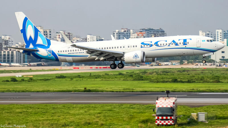 A SCAT Airlines Boeing 737 MAX 8 is landing on a runway with green grass on either side, with buildings visible in the background.