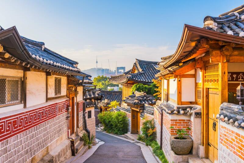 The photo depicts a narrow street in Bukchon Hanok Village in Seoul, South Korea, lined with traditional Korean houses and the Seoul Tower in the background.