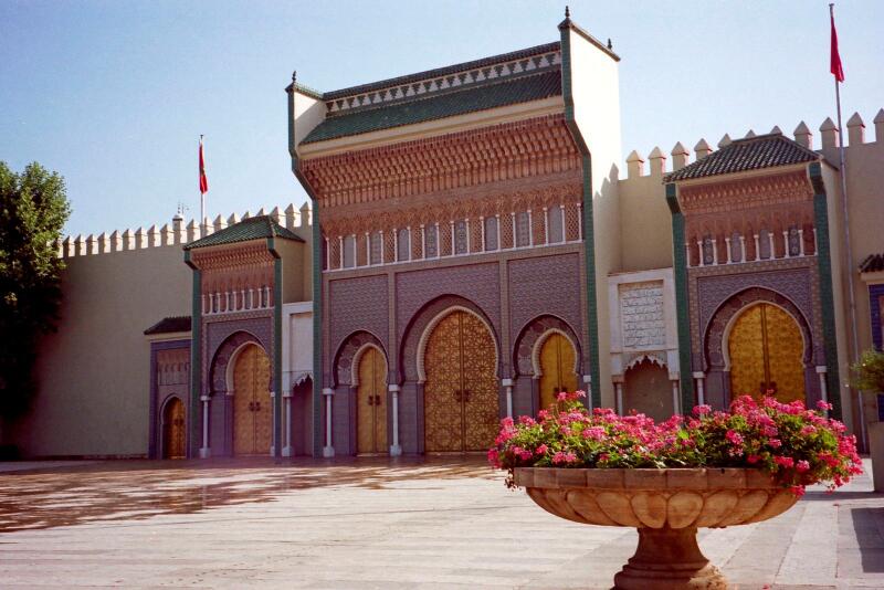 The image shows an ornate building with multiple archways and golden doors, decorated with intricate patterns, and a large stone flower pot with pink flowers in the foreground.
