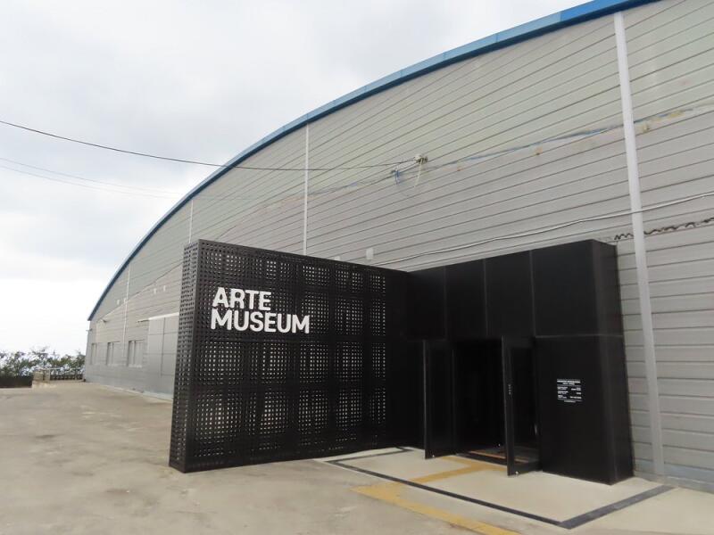 The exterior of the Arte Museum features a gray, barn-like structure with a black, geometric-patterned screen displaying the museum's name in white letters, marking the entrance.