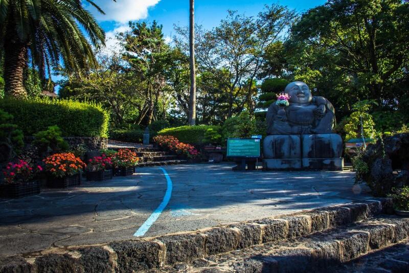 Outdoor shot of a stone Buddha statue holding flowers, located in a garden with trees, flowers, and a blue painted path.