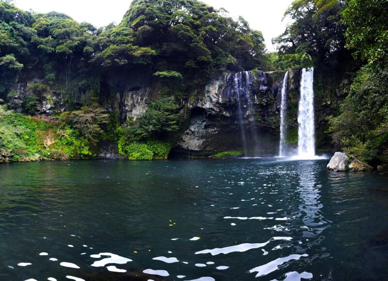 The image shows a scenic view of a waterfall cascading into a dark pool, surrounded by lush greenery and rocky cliffs.