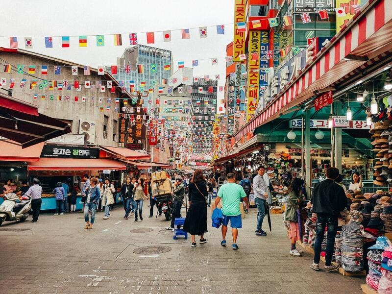 A bustling street scene shows many people walking along a market street lined with shops and food stalls, decorated with numerous international flags overhead.