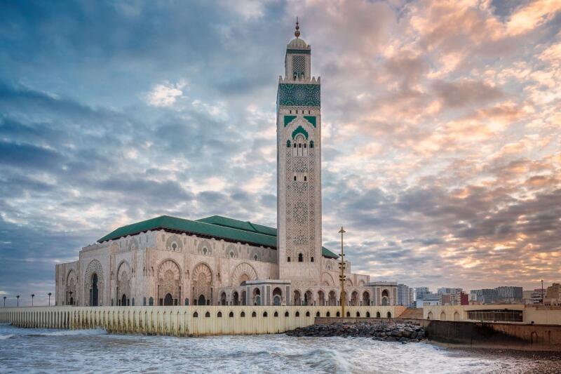 The Hassan II Mosque stands on the coast as waves crash against its base under a cloudy sky.