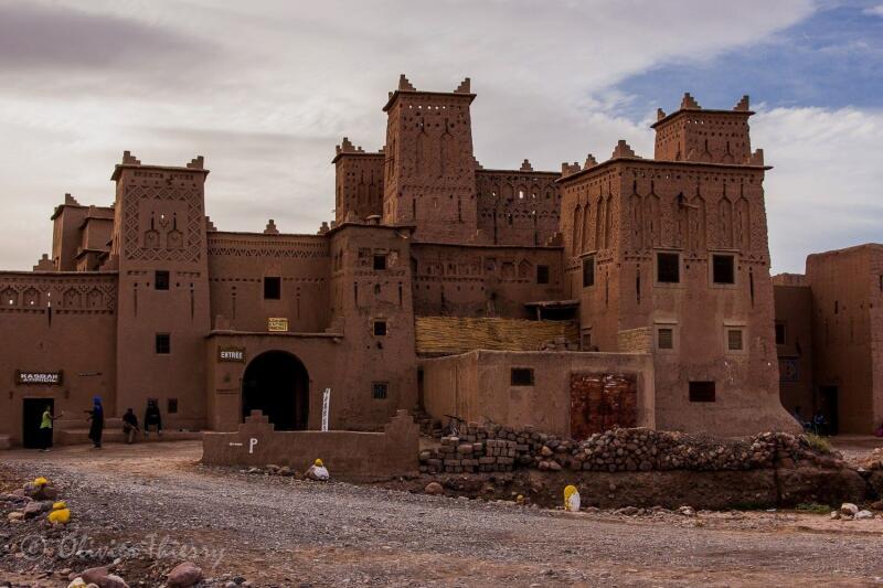 The image shows a kasbah made of mud bricks with several towers and an entrance marked with 'Entree'. People are seen walking around the structure on a rocky terrain under a cloudy sky.