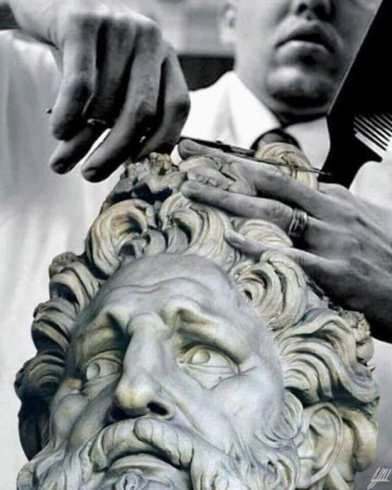 A black and white photo shows a person cutting the hair of a statue head using scissors and a comb.