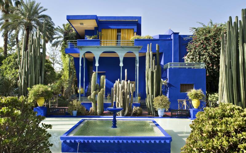 The image shows a vibrant blue building with yellow accents, surrounded by lush greenery and various cacti; in the foreground is a blue fountain.