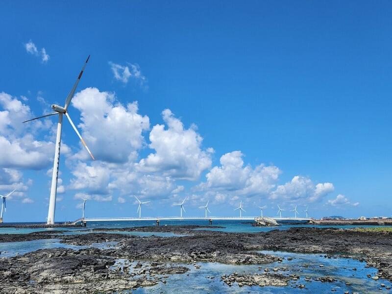 The image shows a landscape with wind turbines, a bridge, and a rocky beach under a blue sky with white clouds. The turbines are tall and white, and the bridge connects them over the water.
