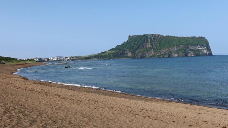 The photo captures a scenic view of a beach with tan sand, a calm blue ocean, and a large, green, flat-topped mountain or hill in the background under a clear blue sky.
