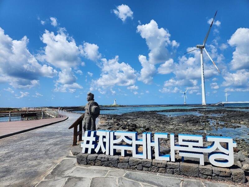 The scenery includes a statue, a sign with the text '#제주바다목장', a stone path, the ocean, and wind turbines under a partly cloudy sky.
