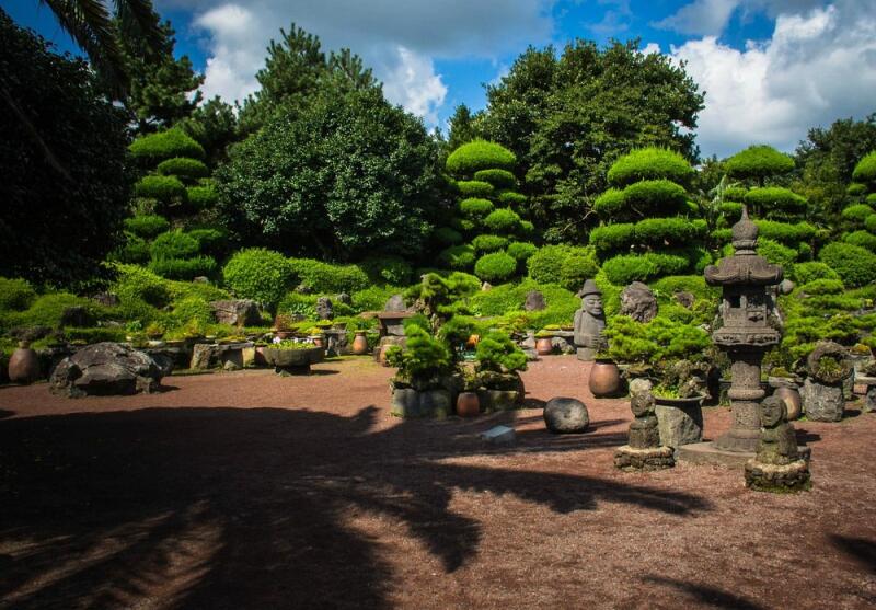 The photo shows a garden with trimmed bushes and trees, stone lanterns, and sculptures on a reddish-brown ground under a partly cloudy sky.