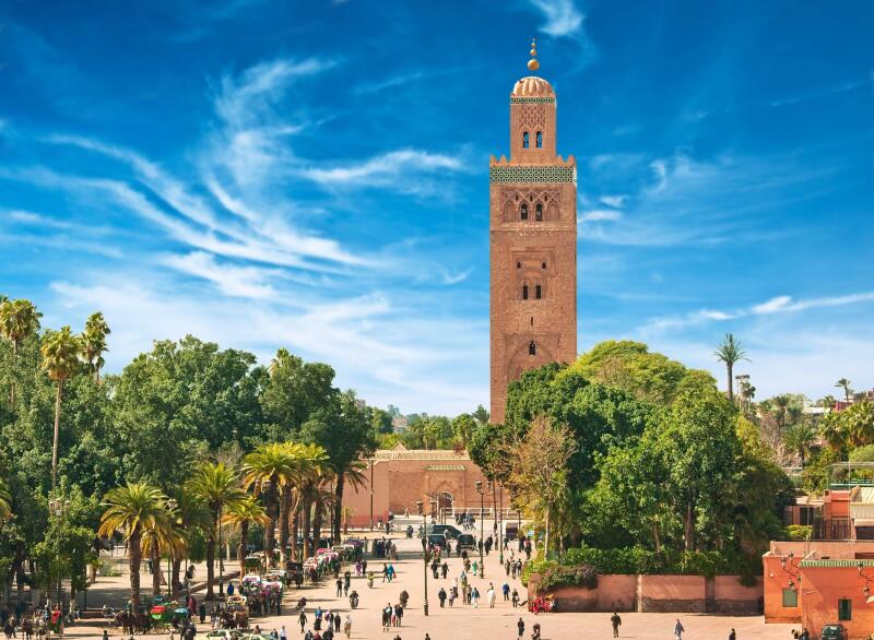 An eye-level shot shows a large, brick-colored mosque tower rising above a square bustling with people and horse-drawn carriages, under a bright blue sky with wispy clouds.