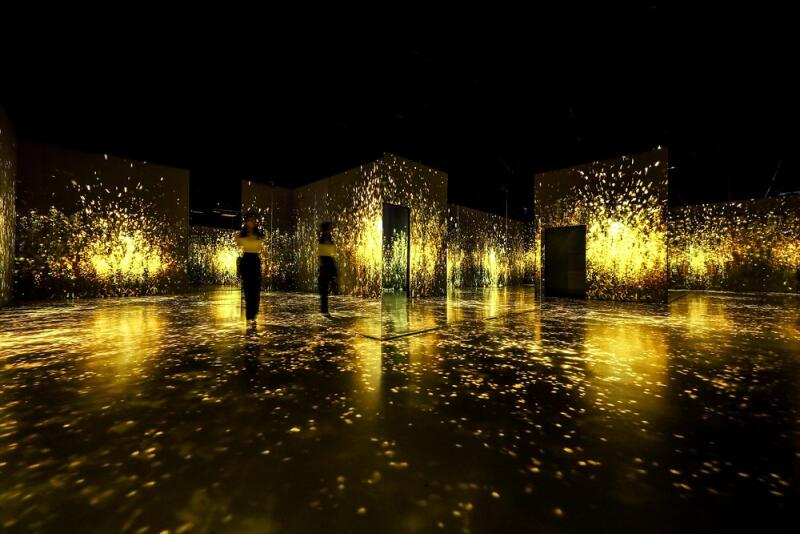 Two people stand in a dark room with walls and floors illuminated with golden light patterns.