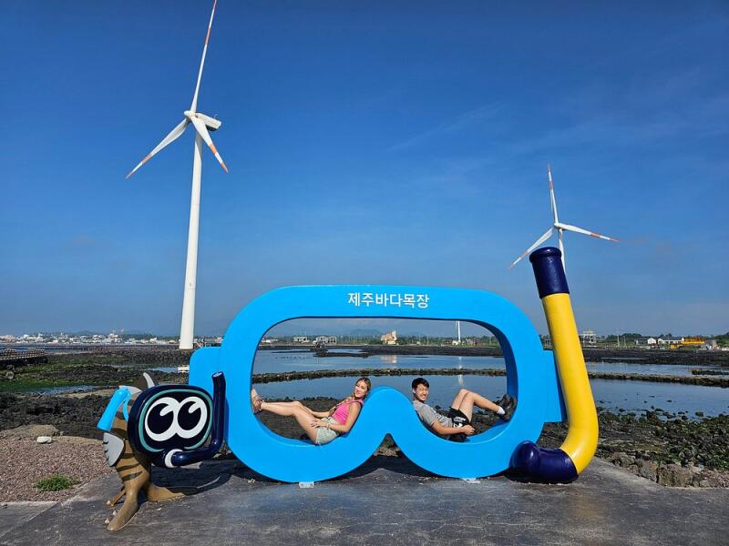 A couple poses within a large, blue snorkeling mask frame with Korean text; a cartoon crab with snorkel gear is on the left, and a large snorkel is on the right, with windmills in the background under a blue sky.