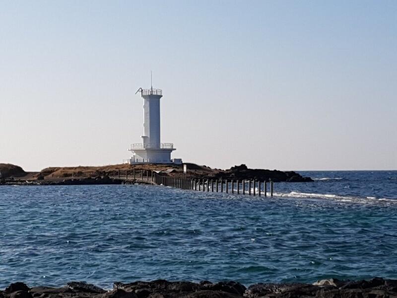 A white lighthouse stands on a small, rocky island, connected to the mainland by a wooden walkway partially submerged in the sea.