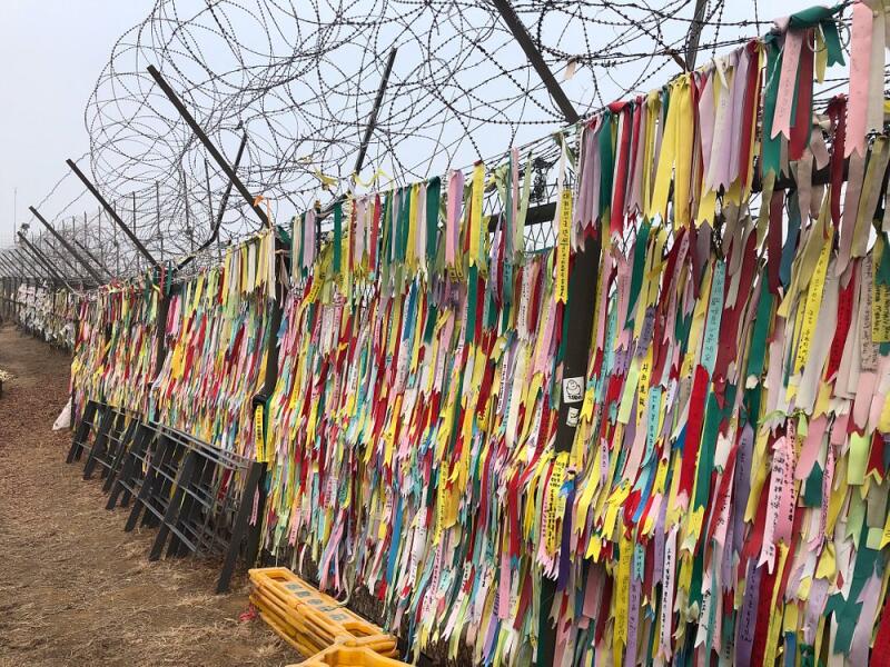 A fence topped with barbed wire is adorned with numerous colorful ribbons bearing written messages.