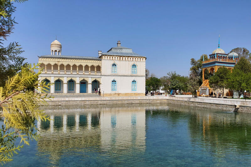 The photo shows a pond reflecting two buildings under a clear blue sky, with one building being white with blue accents and the other being more colorful with yellow and blue.