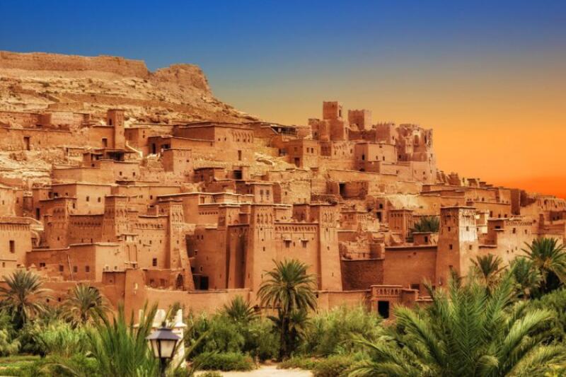 The image features a traditional Moroccan village with brown, mud-brick buildings clustered on a hillside, set against a gradient sky of blue and orange, and framed by palm trees in the foreground.