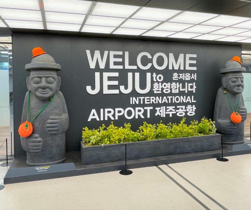 The photo shows the welcome sign at Jeju International Airport, flanked by two Dol Hareubang statues wearing orange hats and carrying pouches. In the center is a planter box with green plants.