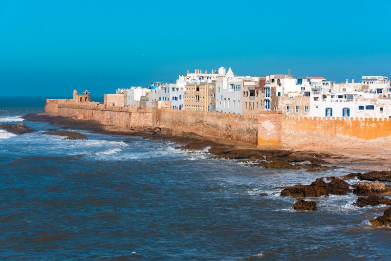 A coastal city featuring a tall, tan stone wall running along the coastline, adjacent to white buildings with blue accents under a bright blue sky and ocean.