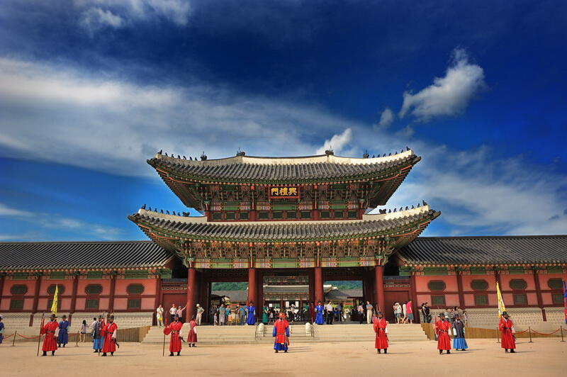 A wide shot of a traditional Korean building with guards in red and blue uniforms standing in front, under a cloudy blue sky. The sign above the gate reads '門樓閣'.