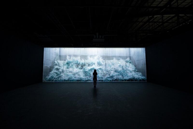 A person stands in a dark room facing a large screen displaying a dynamic image of water, resembling a wave or waterfall.