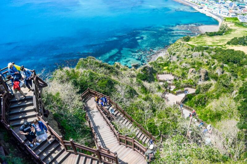 The image shows a coastal landscape with wooden staircases winding down a lush, green hill towards the blue ocean.