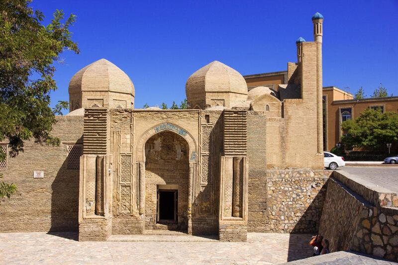 The image shows a tan brick building with two domes and a minaret under a clear blue sky. The entrance features a decorated archway.