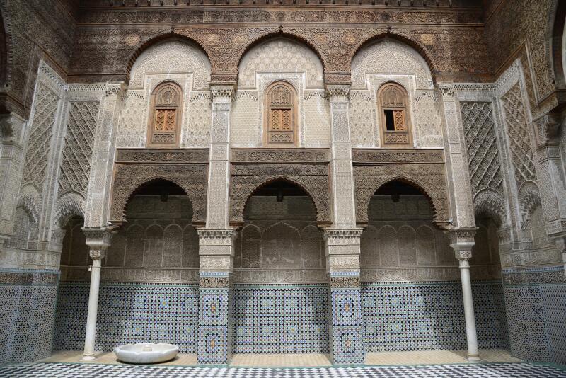 The interior of a building features three arches with carved details, supported by columns, with three windows above the arches, and the lower walls are covered in patterned tiles; a small, round fountain is visible in the foreground on a checkered floor.