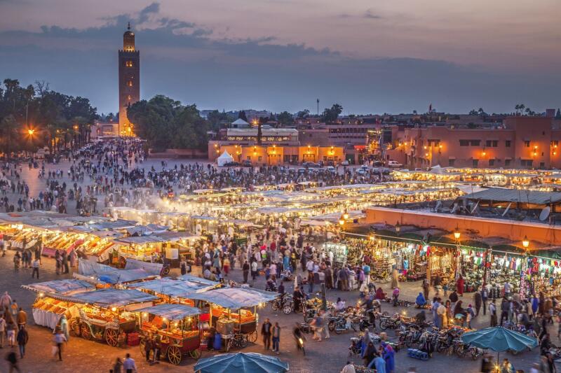 This is an aerial view of Jemaa el-Fna square in Marrakesh, Morocco, at dusk, bustling with people and lit by numerous vendor stalls and building lights, with the Koutoubia Mosque's minaret visible in the background.