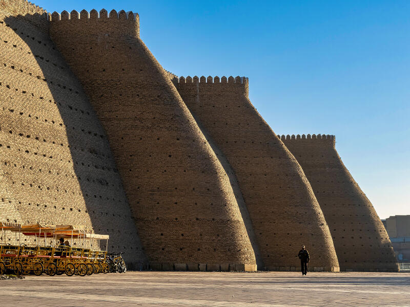 A low-angle shot shows an old brick wall with crenellations, with rows of yellow multi-seated bikes parked nearby and a person walking towards the viewer in a large open area.