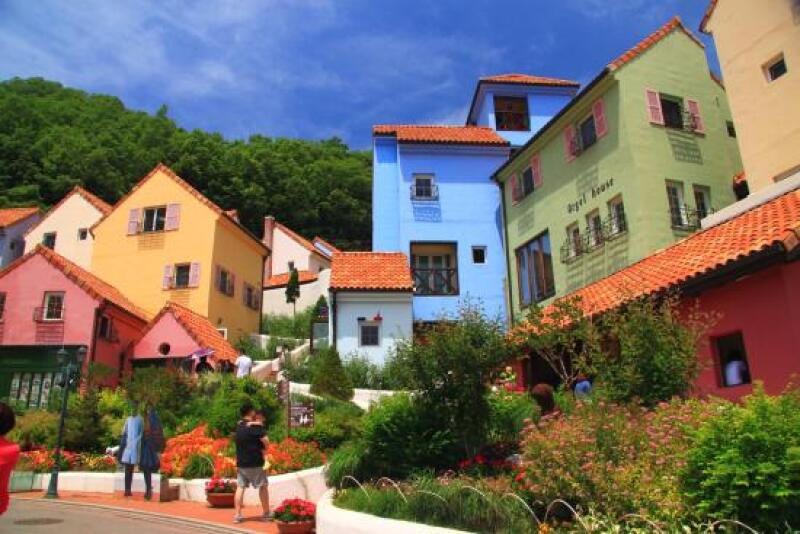 The image shows a colorful village with buildings painted in various shades, including yellow, blue, green, and red, all topped with orange roofs; "Angel House" is visible on the green building, and a lush green hillside forms the backdrop.