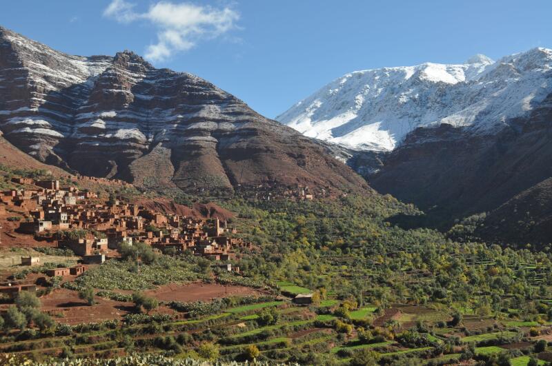 The landscape features a village nestled at the base of layered mountains, with one peak capped in snow, and terraced fields in the foreground under a blue sky.